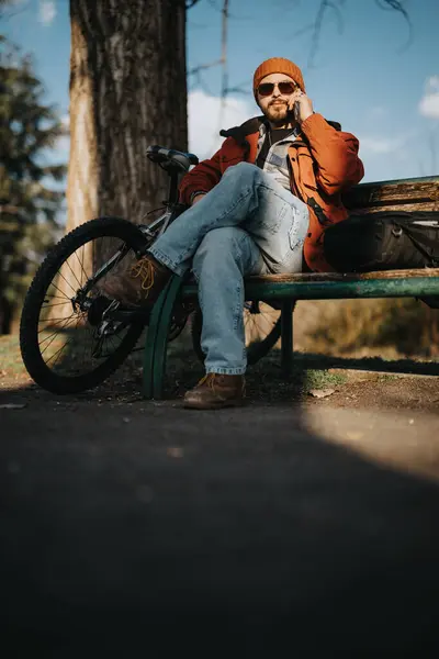 Young casual man sits on a park bench with his bicycle, enjoying a serene, sunny day outdoors.