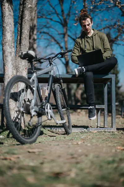 A focused young man with a bun hairstyle sits on a park bench while working on a laptop, his bicycle parked beside him on a sunny day.