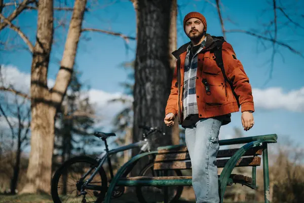 静かな公園の設定で自転車の横に立っているビーニーとオレンジの冬のジャケットのひげ付き男 ロイヤリティフリーのストック画像