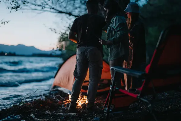 A group of friends gathers around a campfire by a lakeside at night, surrounded by nature, sharing stories and preparing dinner.