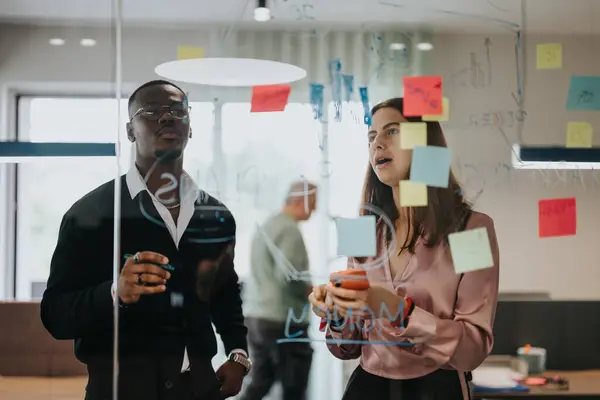 stock image Multiracial young colleagues collaborate in a brainstorming session using sticky notes on a glass wall to map out project ideas and statistics.