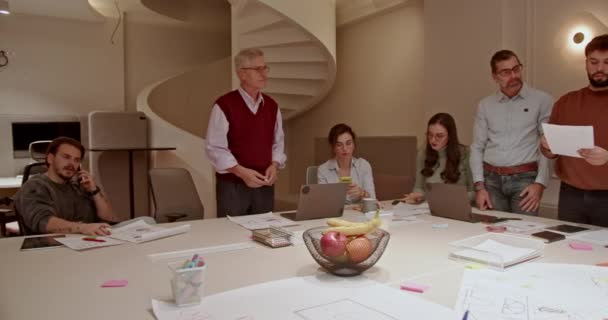 在一个灯火通明的现代化办公室里 一群男女老少的同事围绕着一个堆满文件 手提电脑和一个水果缸的会议桌开展合作 — 图库视频影像
