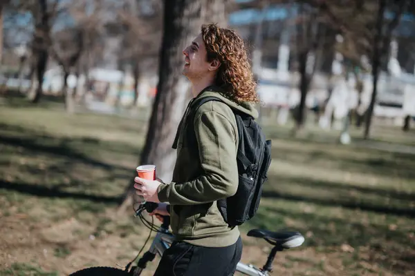 An upbeat casual man paused from biking outdoors, laughing and holding a coffee with a backpack in a serene park setting.