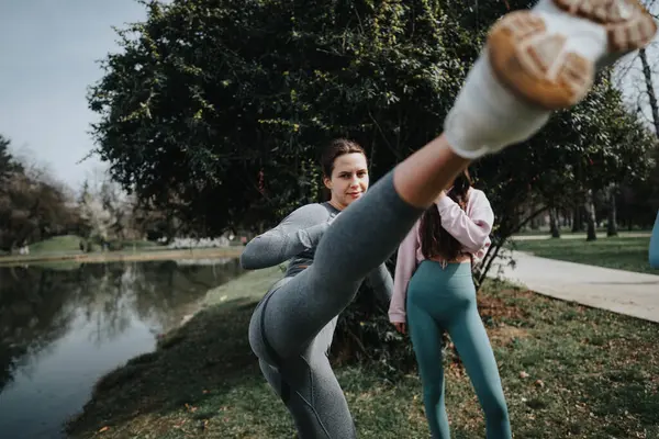 Two women in active wear doing yoga at the park, focusing on a stretch exercise by the lake for mental and physical wellness.