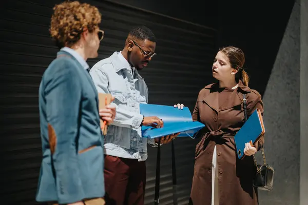 stock image Three diverse business professionals stand outside a city office, engaging in a focused discussion over documents in a casual work meeting.