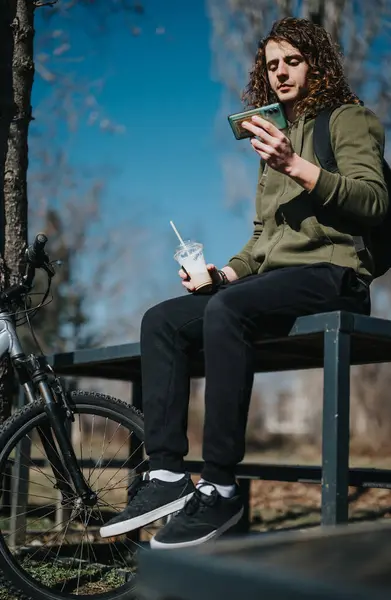 stock image A young curly-haired man dressed in a green hoodie sits on a wooden bench at the park, using his smart phone with a bicycle and a smoothie beside him.