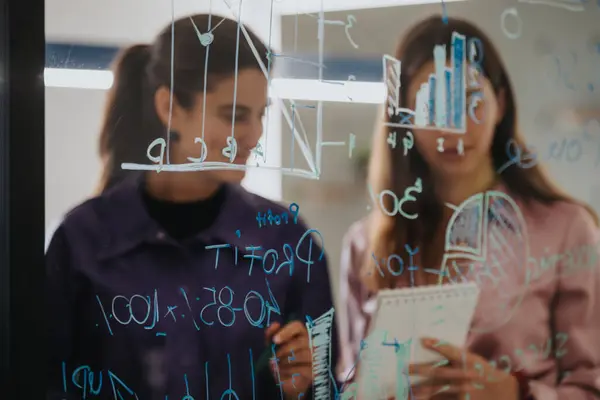 stock image Two businesswomen engage in a strategic session, interpreting and discussing complex statistics written on a transparent glass wall using markers and sticky notes.