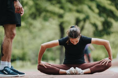 Focused female athlete practices stretching exercises in a park with guidance from a male fitness trainer, surrounded by greenery. clipart