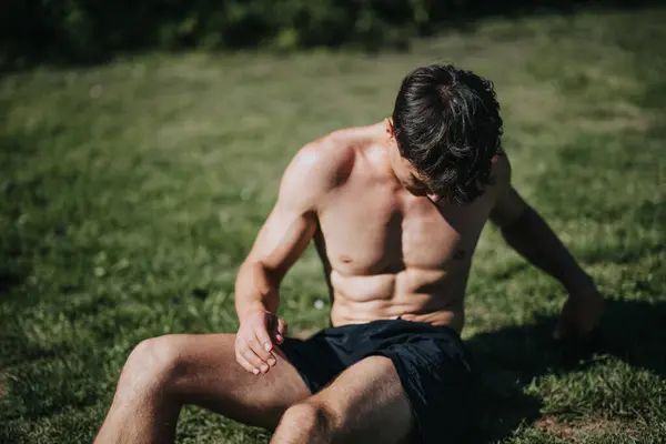 stock image Muscular shirtless man sitting and resting on grass in a park after a rigorous workout under the sun. Fitness and healthy lifestyle concept.