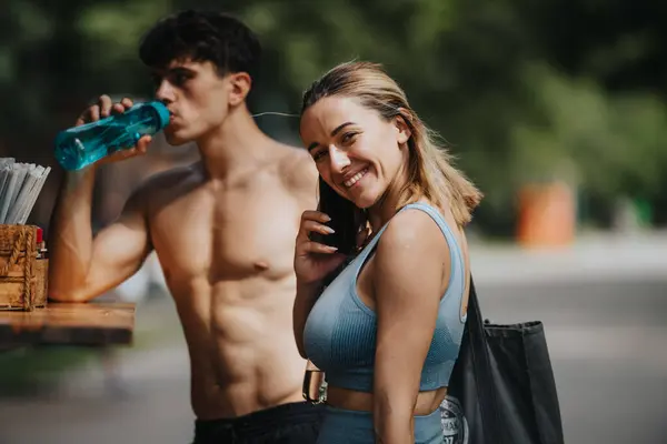 Stock image Happy sports couple enjoy refreshing drinks after a workout outdoors. Shirtless man drinking water and woman smiling with phone call.