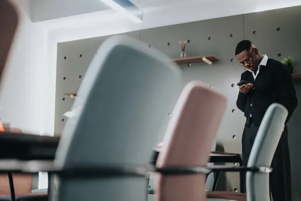 Stock image An African American male in business attire attentively using his smart phone amidst contemporary office furniture. Capturing a moment of technology in business.