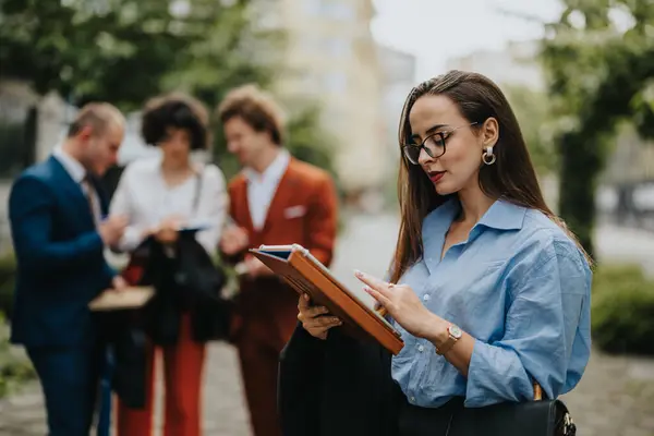 stock image Businesswoman using a tablet outdoors while colleagues interact in the background during an informal team meeting. Concept of teamwork, technology, and outdoor working environment.