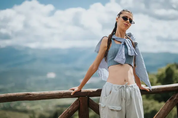 stock image Stylish young woman with braided hair and sunglasses posing against a picturesque mountain backdrop. She exudes confidence and summer fashion vibes.