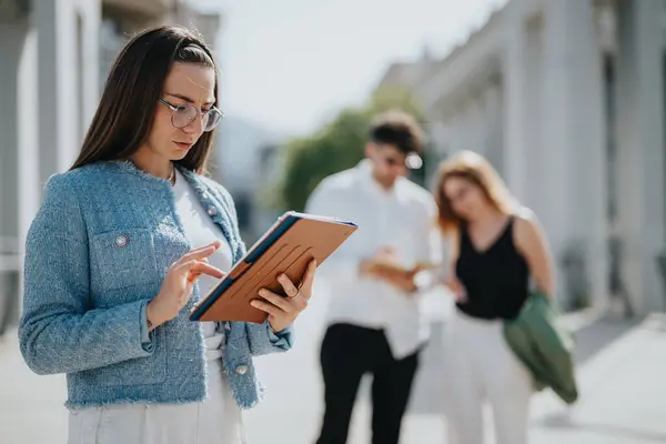 stock image Businesswoman focused on tablet with colleagues discussing in the background. Outdoor business interaction with technology and teamwork.