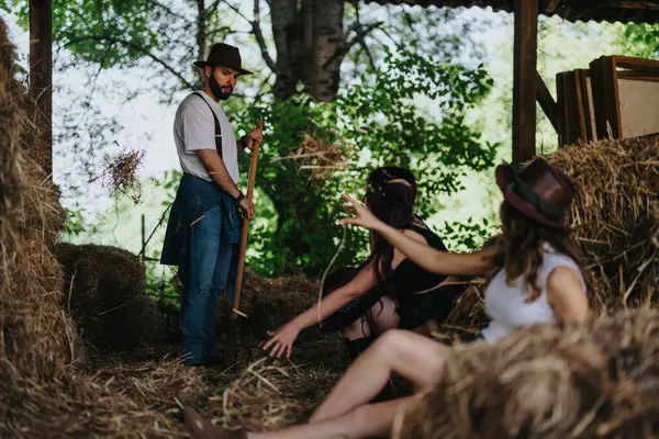 stock image Group of friends in rustic setting, one holding farm tool while others relax on hay bales with greenery in background