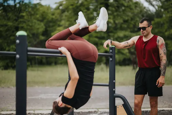 stock image Outdoor fitness session capturing a woman doing an inverted exercise with assistance from a male coach, both in athletic wear in a park setting.