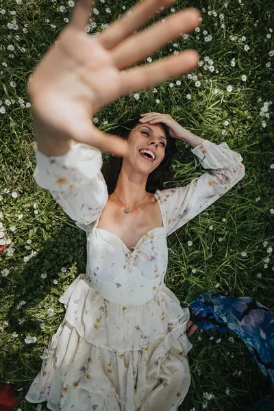 stock image A smiling girl in a floral dress stretches her hand towards the camera while lying on a grassy field dotted with daisies, expressing joy and spontaneity.