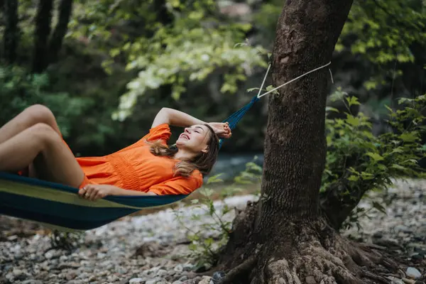 stock image Young woman wearing an orange dress relaxing in a hammock in a forest. She is smiling and enjoying the peaceful natural surroundings.