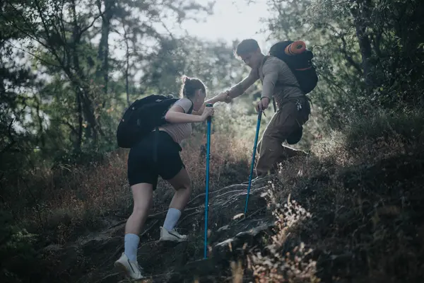 stock image Two hikers helping each other ascend a steep trail in a forest, showcasing teamwork and outdoor adventure.