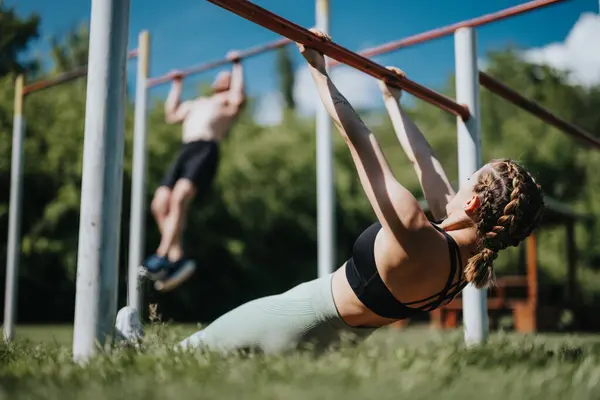 stock image Man and woman exercising together on fitness bars in a sunny park. Engaging in strength training and outdoor fitness activities.