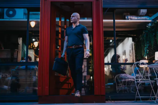 stock image A confident businessman in casual attire holding a briefcase walks out of a modern building at night, exuding professionalism