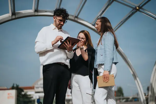 Stock image Three business colleagues collaborating on a project using a tablet, with bright blue sky and modern architecture in the background.