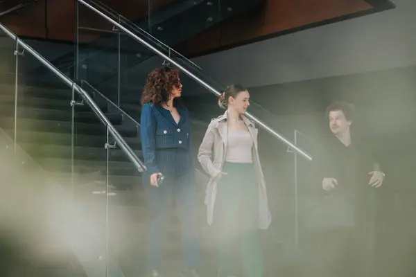 stock image Three businesspeople walking down stairs in a modern office, engaged in conversation and dressed in professional attire.