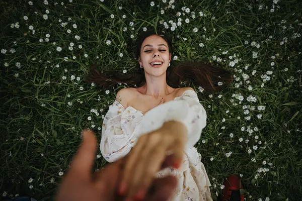 Stock image A joyful moment captured as a young girl lies on a grassy field, surrounded by small daisies, reaching out to the camera in a playful gesture.