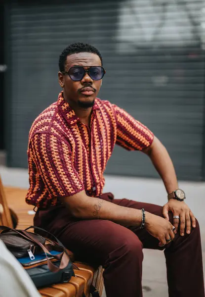 stock image Confident man wearing sunglasses and a fashionable shirt, sitting on a bench outdoors, exuding style and cool demeanor.