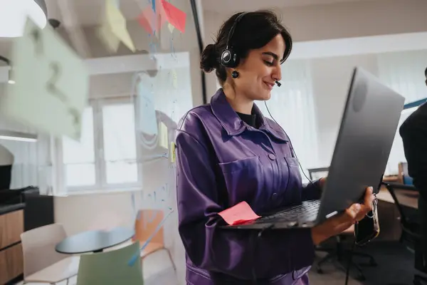 stock image An Indian, female employee in a vibrant purple outfit joyfully engages in her work on a laptop, equipped with a headset in a modern office setting.