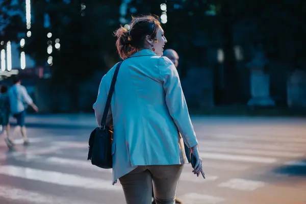 stock image Back view of a businesswoman walking in the city at night, holding a phone and carrying a shoulder bag.