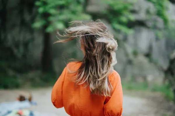 stock image Back view of a person with long hair in an orange shirt enjoying a windy day outdoors, surrounded by nature.