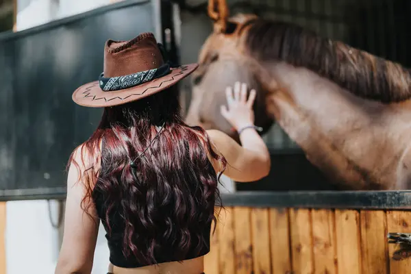 stock image Woman with long wavy hair and cowboy hat gently petting a horse in a stable. A peaceful, heartfelt moment of connection between human and animal.