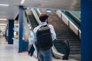 Rear view of a businessman with a backpack and notepad walking through a modern airport terminal towards escalators. clipart