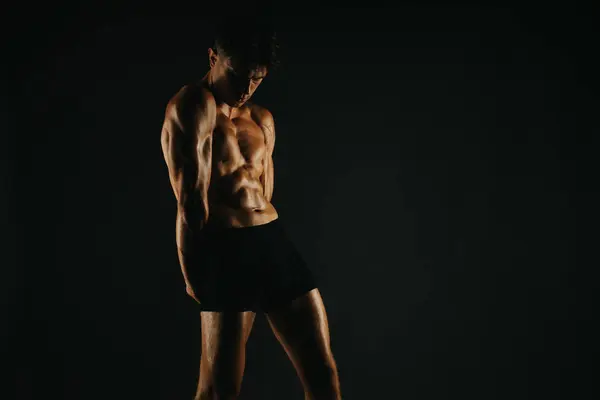 stock image Fit male model displaying his well-defined abs and muscular arms. Posing confidently in a dark setting highlighting his fitness.