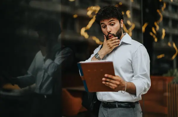 stock image Young businessman with surprised expression examining a document outside. City reflections visible on glass. Concept of unexpected news, urban business life.