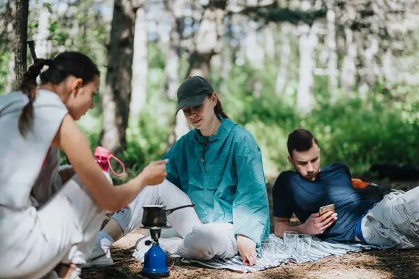 stock image Group of friends enjoying a camping trip in the forest, sitting on a blanket, preparing coffee, and relaxing on a sunny day.