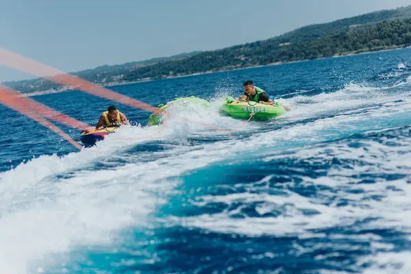 stock image Two friends enjoying an exhilarating speedboat tubing experience on a bright summer day in the sea, creating waves and excitement.