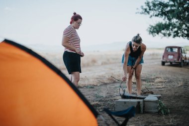 Two friends setting up a campfire during a camping adventure in the mountains. Enjoying nature and relaxation. clipart