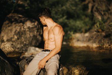 A shirtless man sits on rocks by water, basking in natural sunlight in an outdoor setting, conveying ruggedness and fitness. clipart