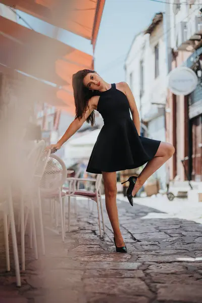 stock image Young woman in black dress striking a playful pose on a charming cobblestone street, blending elegance and .