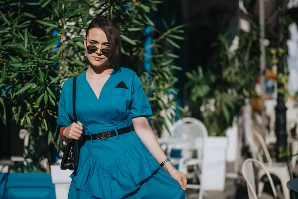 stock image Young woman in blue dress and sunglasses enjoying a sunny day outside a trendy cafe surrounded by greenery.