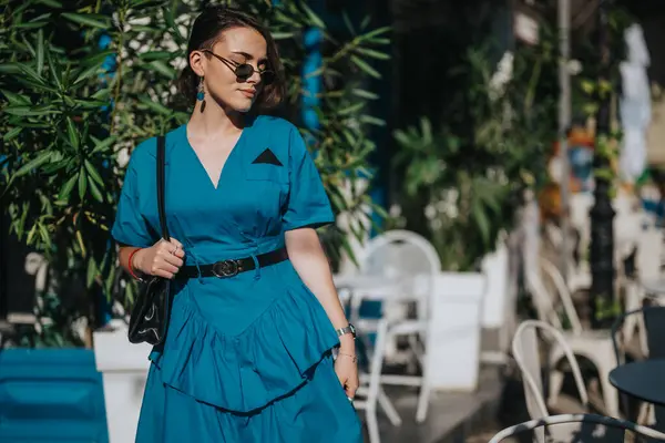 Stock image Fashionable woman wearing a blue dress and sunglasses, striking a pose while enjoying a sunny day. Urban outdoor setting with greenery and casual seating.