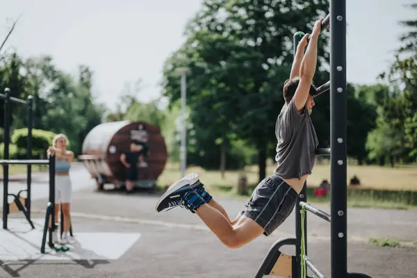 stock image Young man performing calisthenics workout on bar in a sunny park while friends watch. Outdoor exercise and fitness training