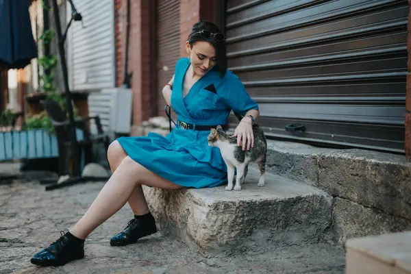 stock image Young woman in blue dress sitting outdoors, petting a friendly tabby cat on a stone step in an urban environment.