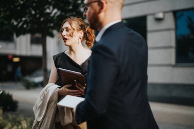 Business professionals discussing work while walking on a city street, holding notebooks and documents, in a casual yet professional atmosphere. clipart