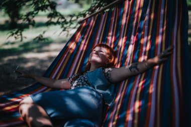 Woman lying on a colorful hammock outdoors, surrounded by nature, enjoying a serene and tranquil moment. clipart