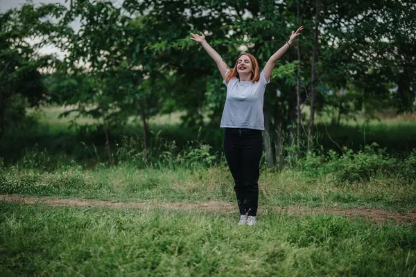 stock image A happy woman stands with arms raised, enjoying the fresh air in a vibrant green park setting.