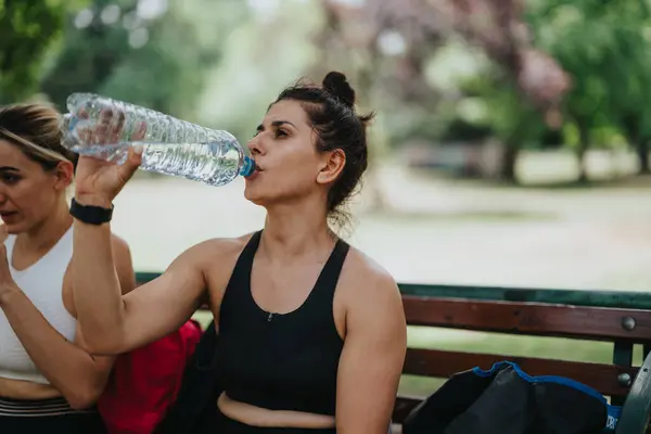 stock image Two women taking a break and hydrating after exercise, sitting on a park bench on a sunny day.