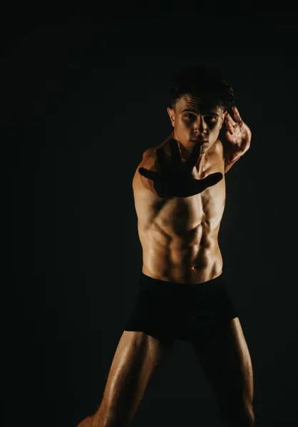 Stock image Young muscular man striking a powerful martial arts pose during a workout session, showcasing strength and determination in dramatic lighting against a dark background.